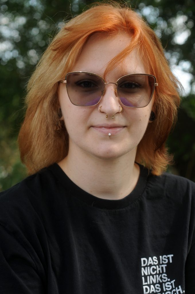Foto von Niculina Pruscini, einer weißen Person mit langen rötlichen Haaren, Sonnenbrille, Piercings in Mund und Nase und einem schwarzen T-Shirt mit der Aufschrift Das ist nicht links, das ist logisch.